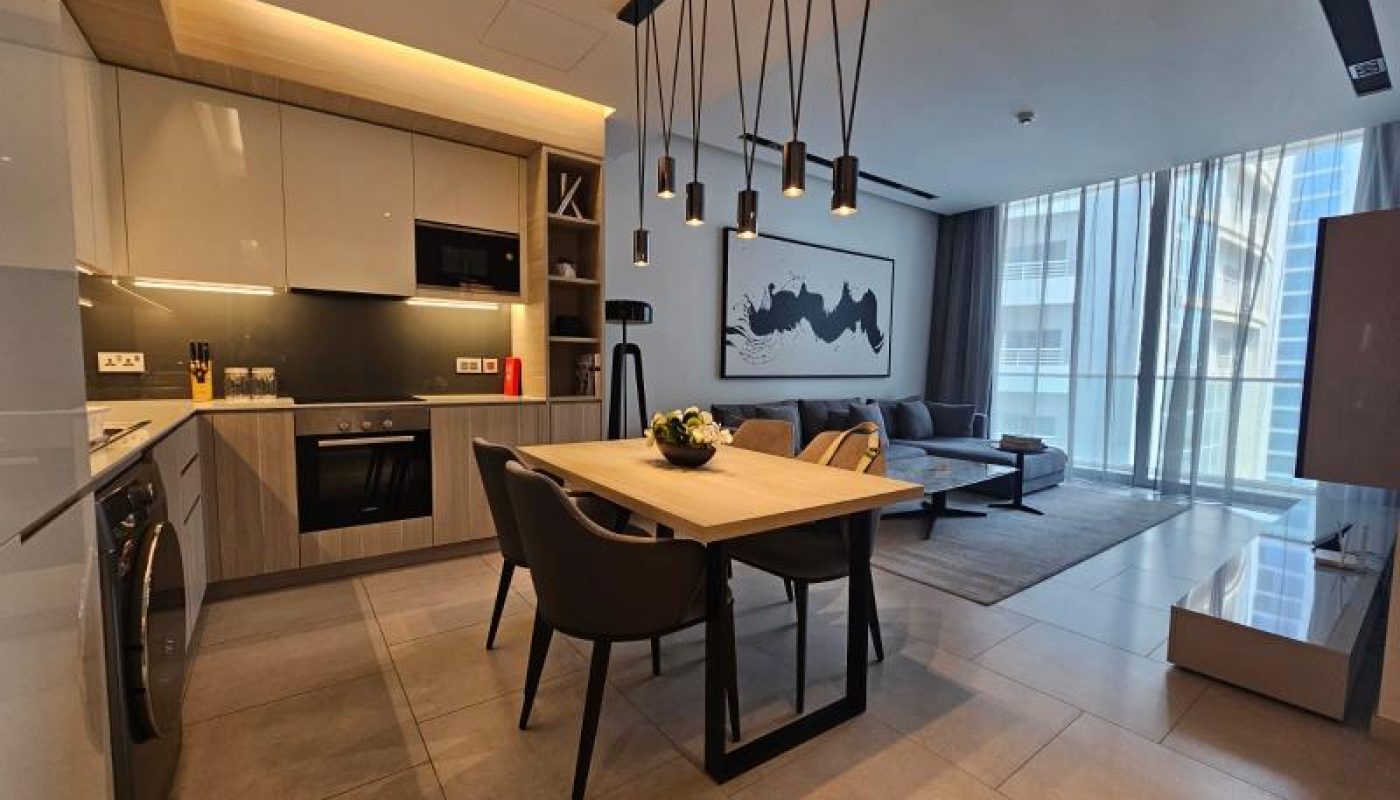 Modern open-plan apartment with a kitchen, dining table, and living area featuring contemporary furnishings and large windows with sheer curtains.