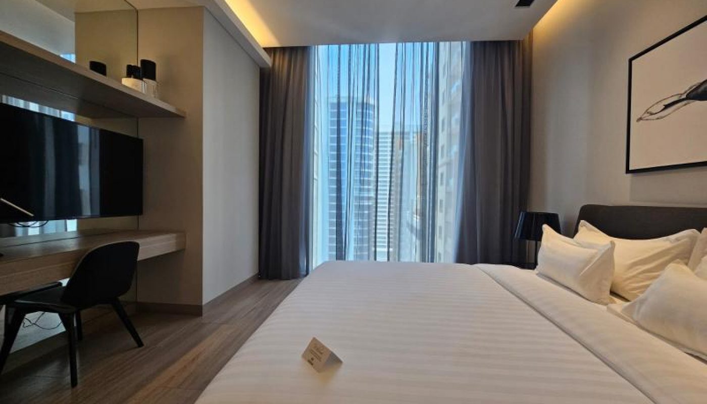 Modern hotel room with neatly made bed, desk and chair, wall-mounted TV, large window with city view, and neutral decor.