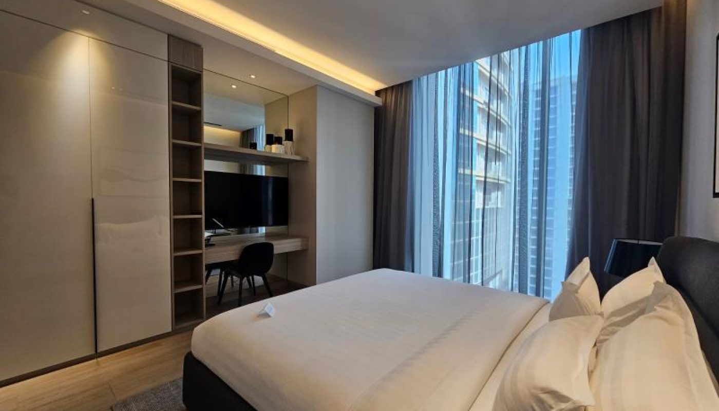 Modern hotel room with a neatly made double bed, desk and chair, large wardrobe, and floor-to-ceiling windows with cityscape views.