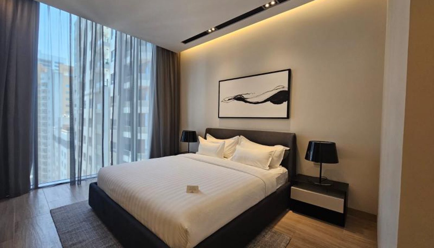 A modern hotel room with a large bed, two bedside tables with lamps, a rug, wall art above the bed, and floor-to-ceiling windows with city views.