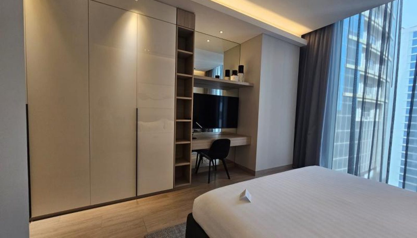 Modern hotel room with a large bed, built-in wardrobe, desk with chair, wall-mounted TV, and floor-to-ceiling windows showing city buildings outside.