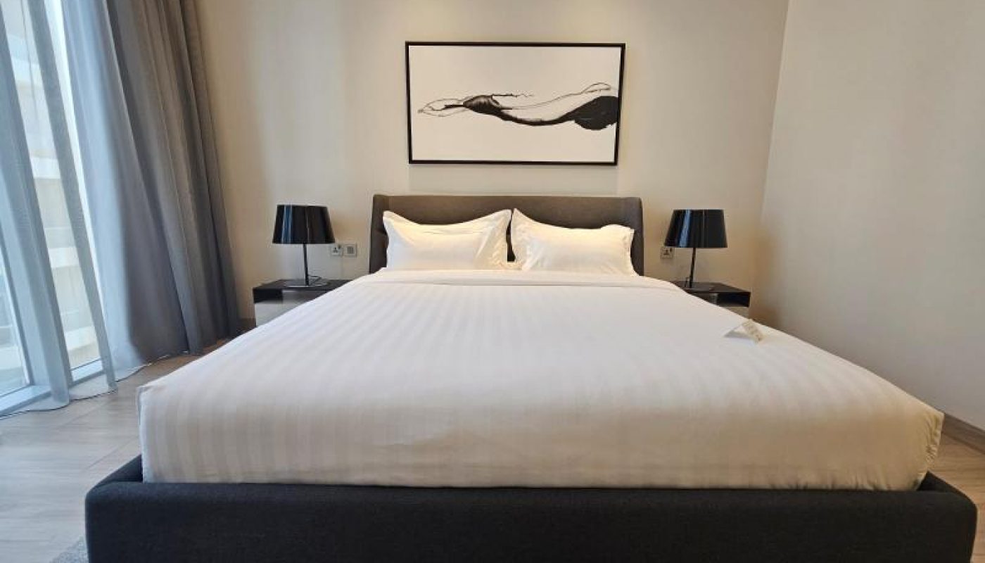 A neatly made bed with white linens is centered between two black lamps and side tables, with a minimalist abstract artwork hanging above the headboard.