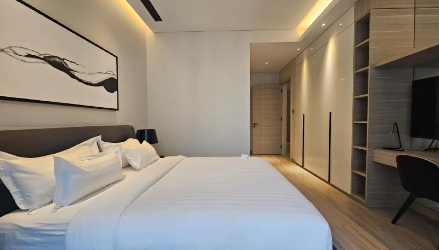 Modern bedroom with a neatly made bed, a large abstract artwork above the headboard, built-in wardrobes, and a desk with a chair on the right side.