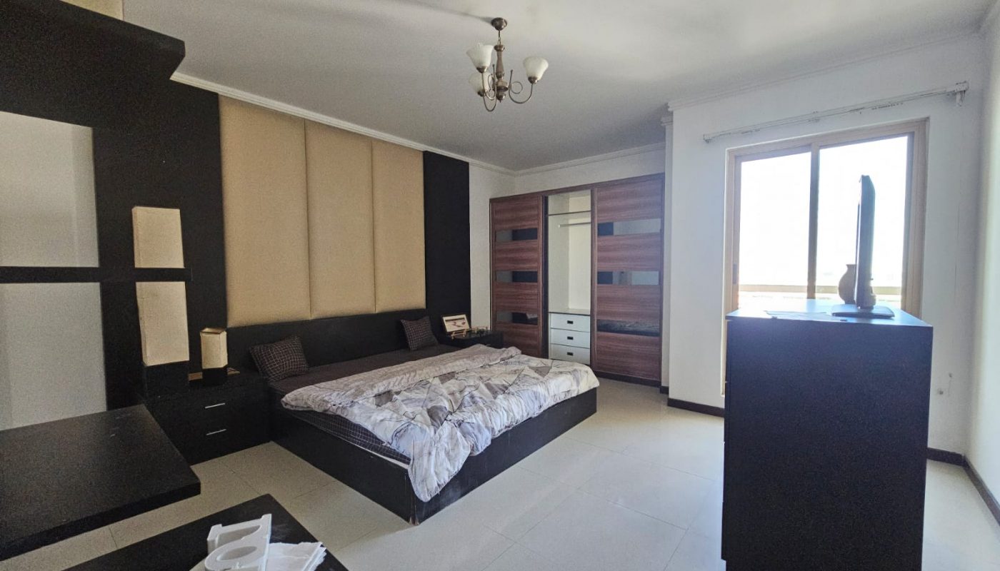 Modern bedroom with a large bed, built-in wardrobe, dresser with a TV, and a window letting in natural light. Neutral color palette with black, beige, and wood tones.