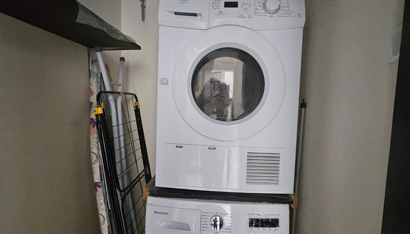 A stacked washer and dryer in a small laundry room with shelves above, an ironing board, drying rack, and a white trash bin on the floor.
