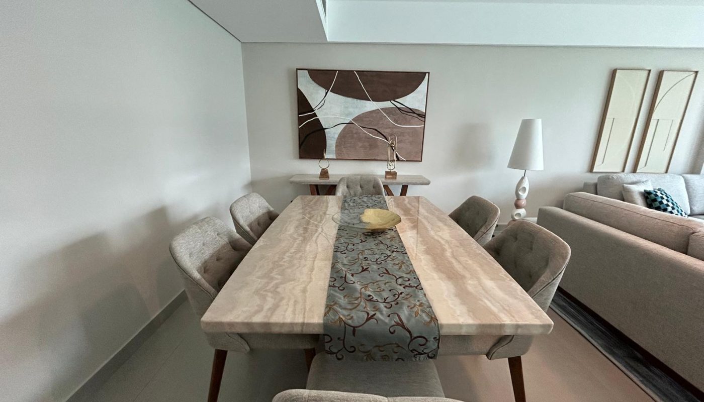 Rectangular marble dining table with six upholstered chairs, a decorative bowl, abstract wall art, lamp, and a beige sofa visible in a modern living room.
