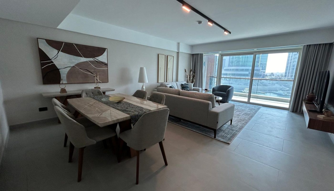 A modern living and dining area with neutral tones, featuring a dining table, sofas, wall art, and large windows overlooking a cityscape.