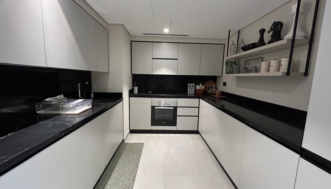Modern kitchen with white cabinets, black countertops, built-in oven, open shelves holding decor items, and a drying rack beside the sink. The floor is light-colored tile.