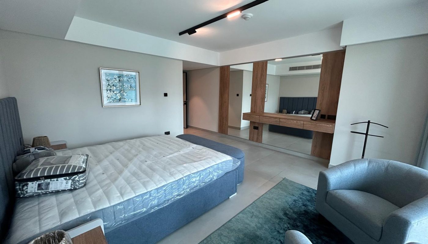 Modern bedroom with a large bed, gray upholstered headboard, blue rug, armchairs, wooden accents, wall art, and a wide mirror above a wooden desk.