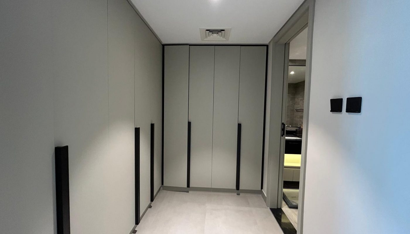 A modern, minimalist hallway with built-in grey wardrobes, black handles, and light-colored flooring, leading to a room visible through an open door.