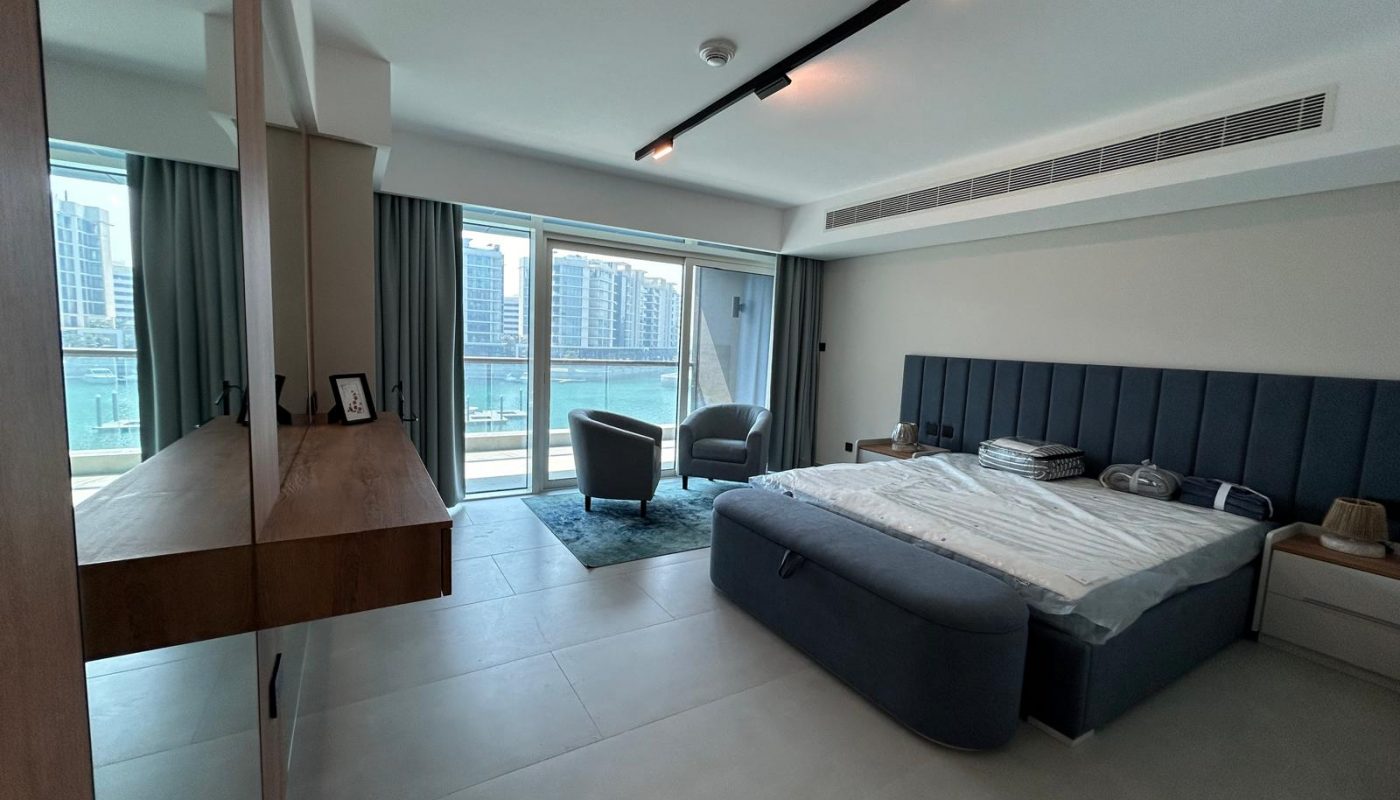 Spacious modern bedroom with large windows, a blue upholstered bed, two chairs by a small table, and a view of high-rise buildings and water outside.