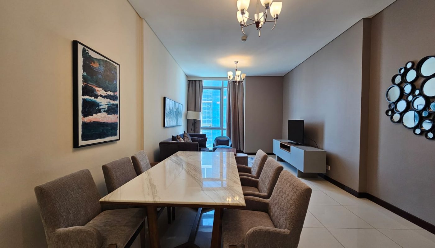 A modern dining and living area with a marble table, six chairs, wall art, a TV, and large windows letting in natural light.