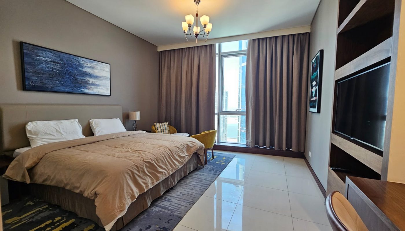 Modern hotel room with a large bed, armchair, artwork, TV on a wall shelf, desk, and floor-to-ceiling window with closed curtains.