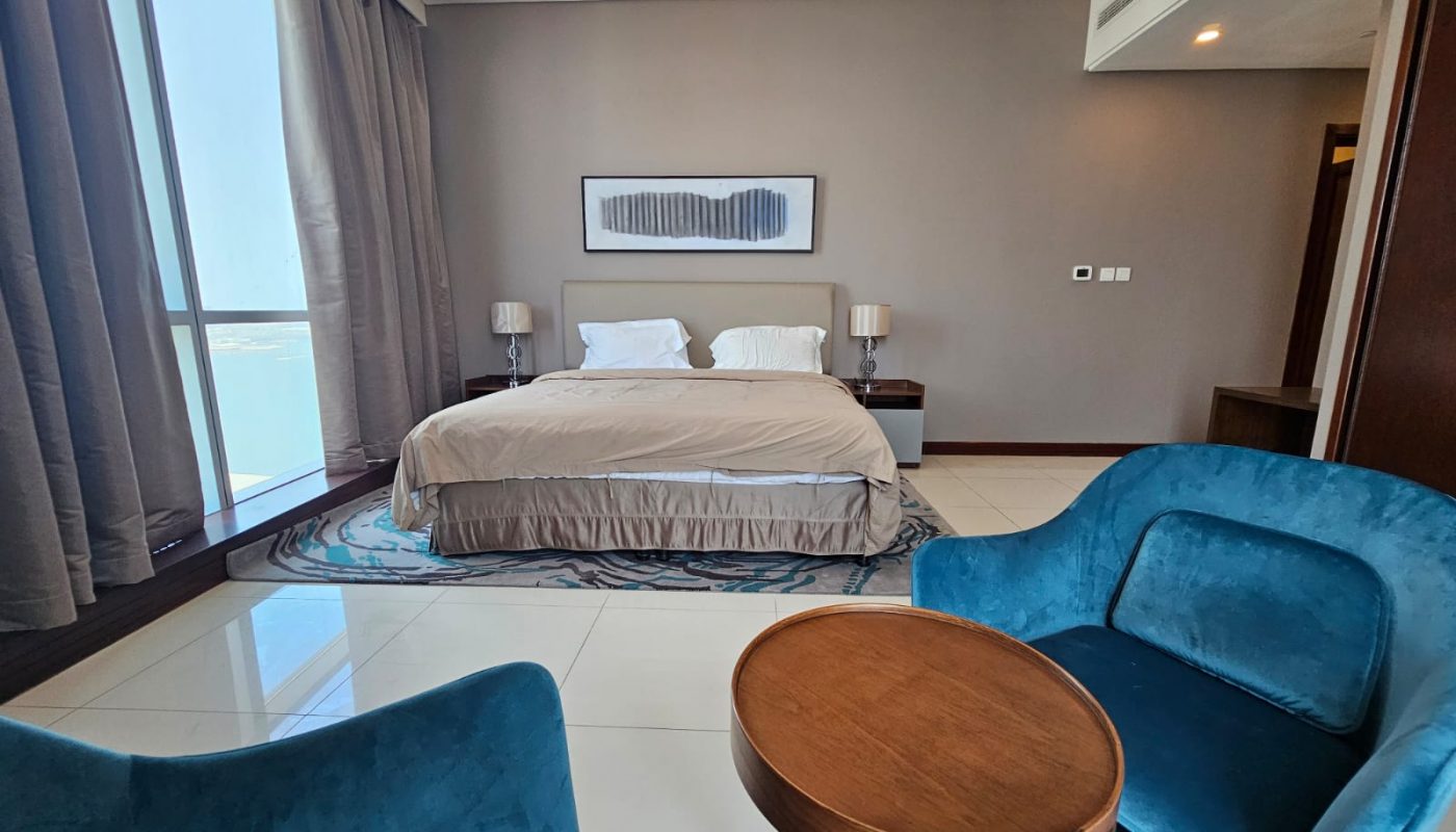 A modern hotel room with a double bed, two bedside tables with lamps, a large window with curtains, two blue chairs, and a round wooden table.