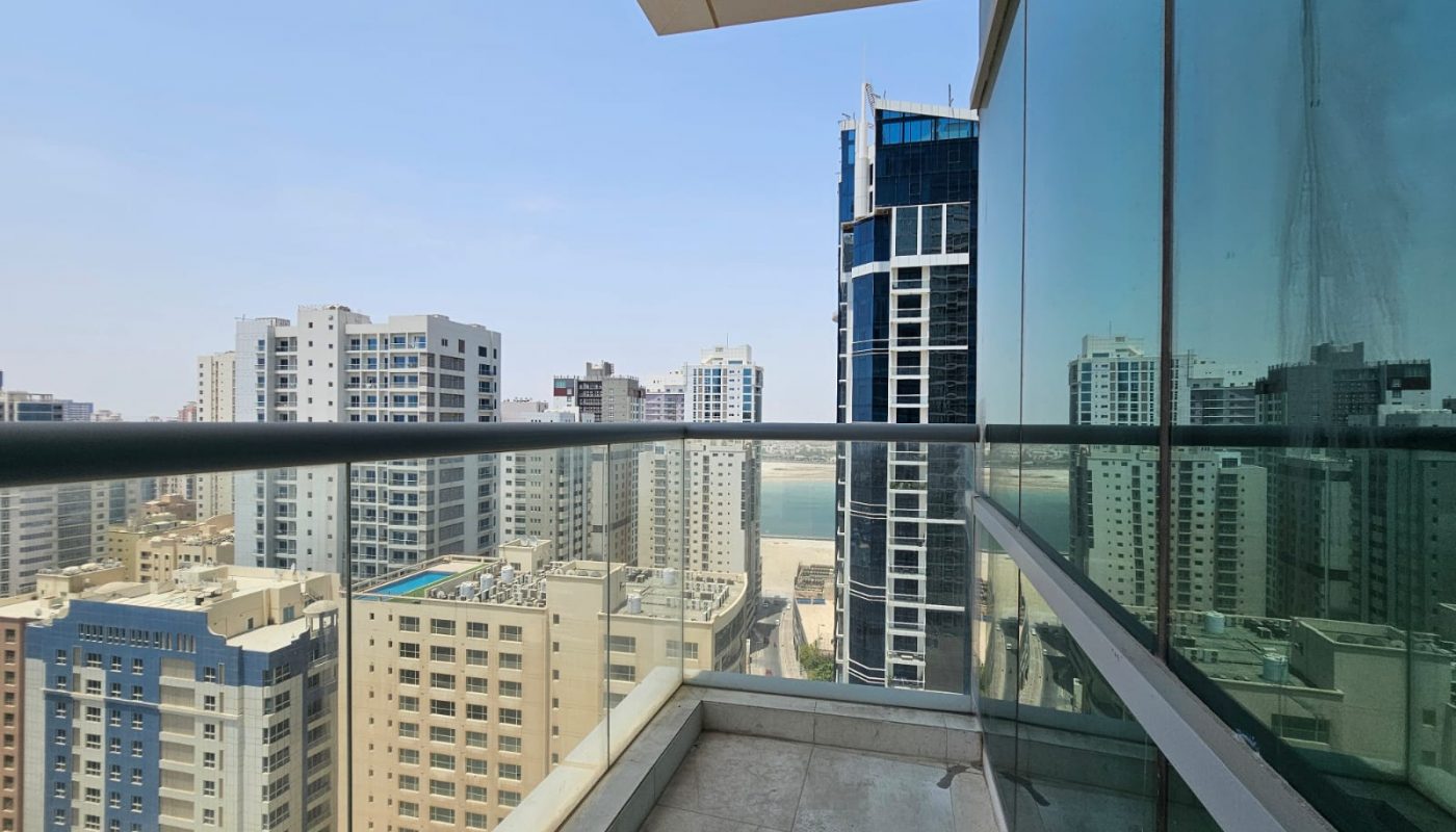 View from a high-rise balcony overlooking modern city buildings with a partial glass reflection and a distant body of water under a clear sky.