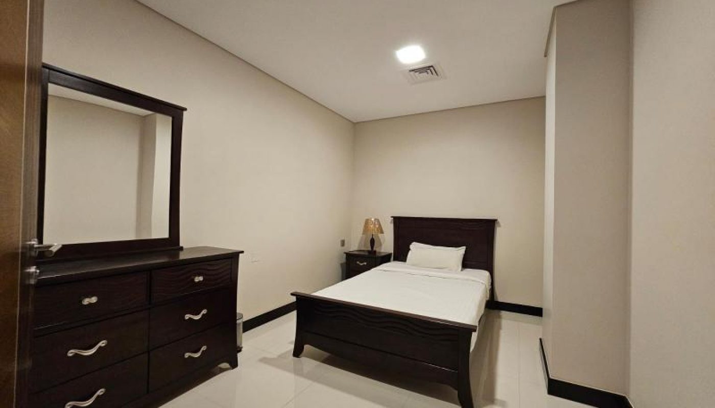 A simple bedroom with a single bed, a wooden dresser with a mirror, a bedside table with a lamp, and cream-colored walls and tiles.
