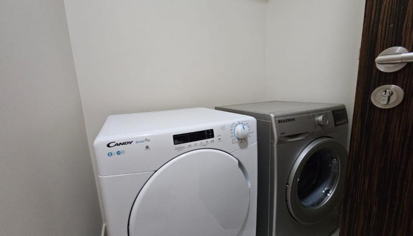 A white Candy washing machine and a silver Electrolux dryer are placed side by side in a small laundry room with white walls.