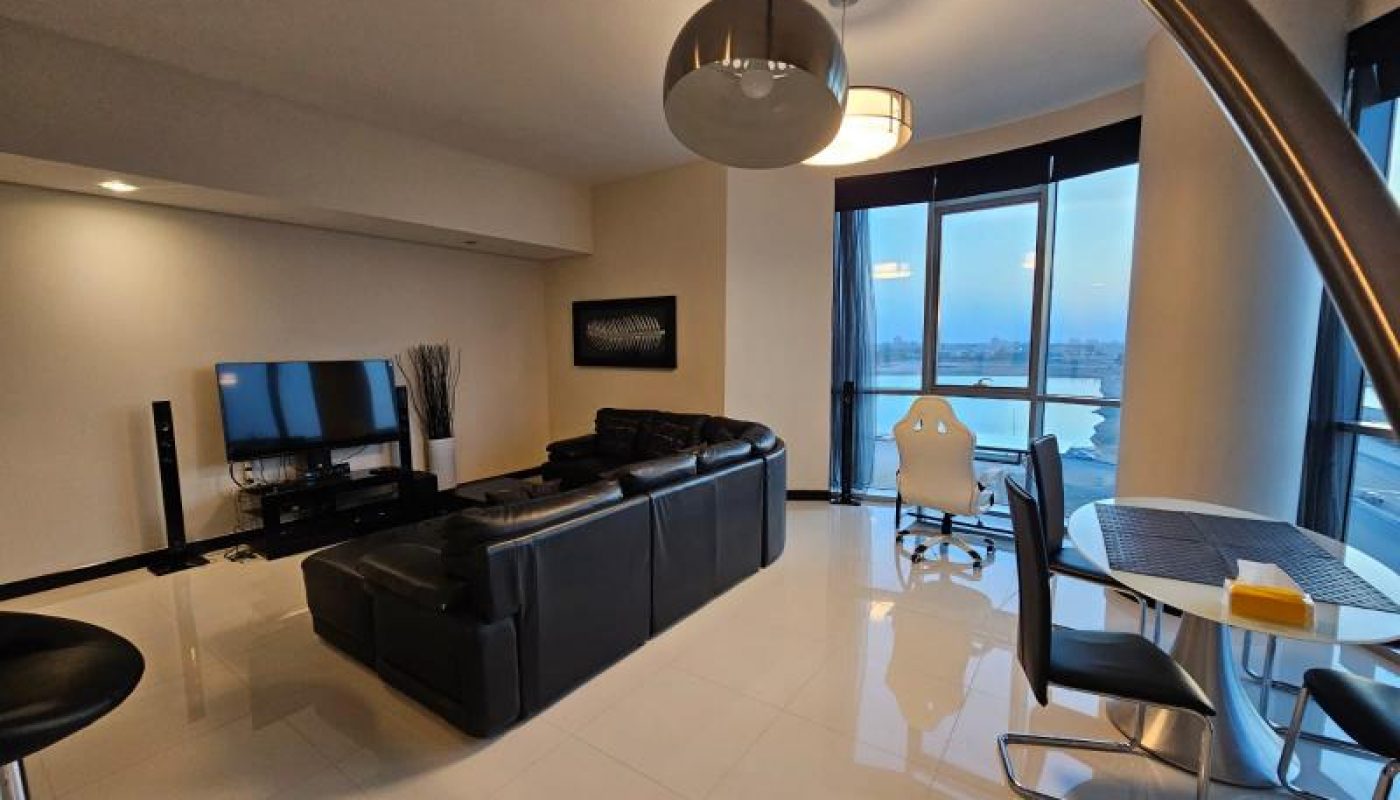 Modern living room with black leather sectional sofa, TV, desk with white chair by large windows, and a dining table with chairs on a tiled floor.