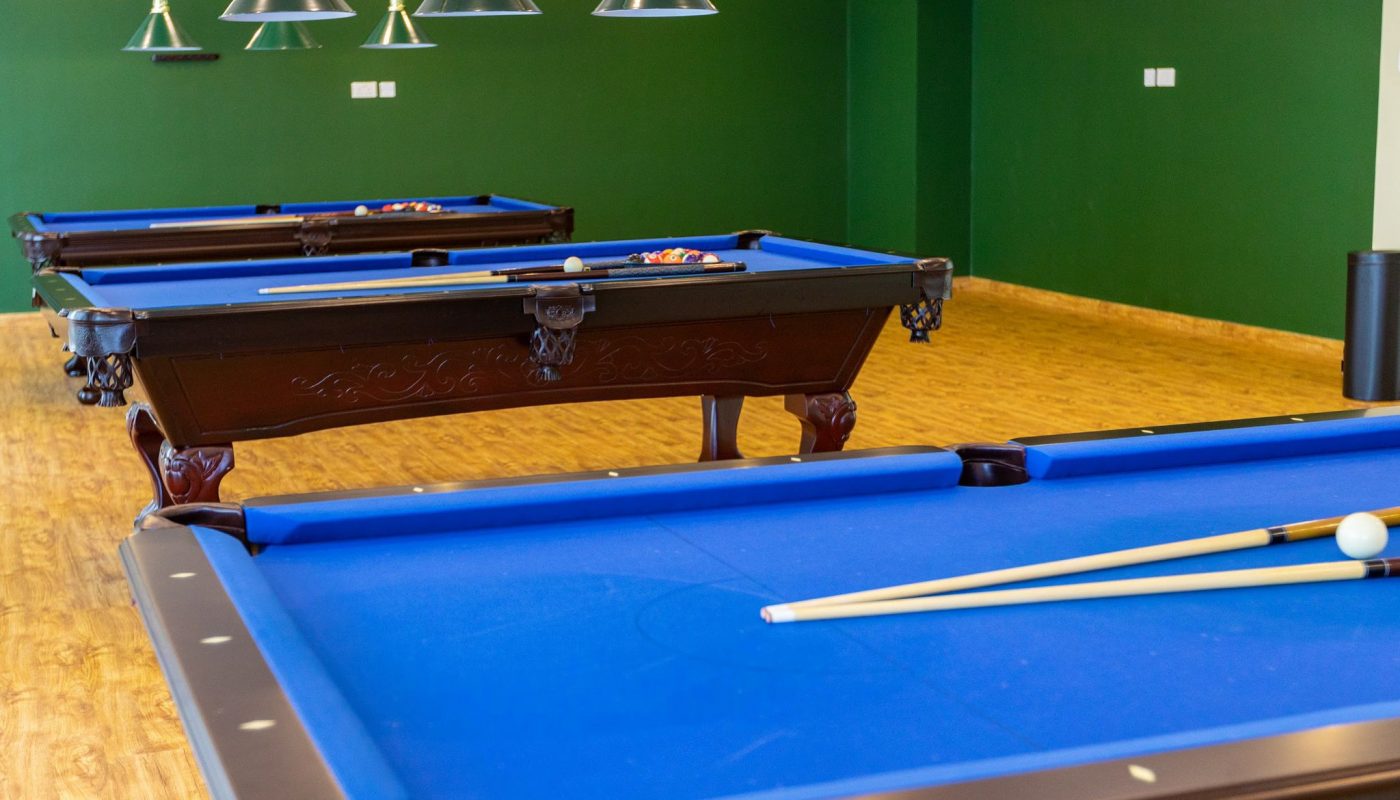 Three pool tables with blue felt, pool cues, and balls, set in a room with green walls, wooden floor, and overhead lighting.