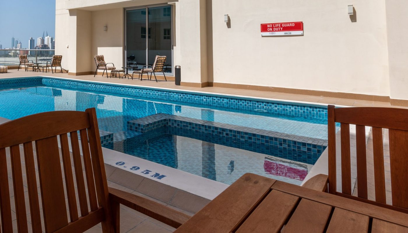 Outdoor swimming pool with blue tiles, wooden tables and chairs in the foreground, and a "No Lifeguard on Duty" sign on the back wall. The pool depth is marked as 0.95 meters.