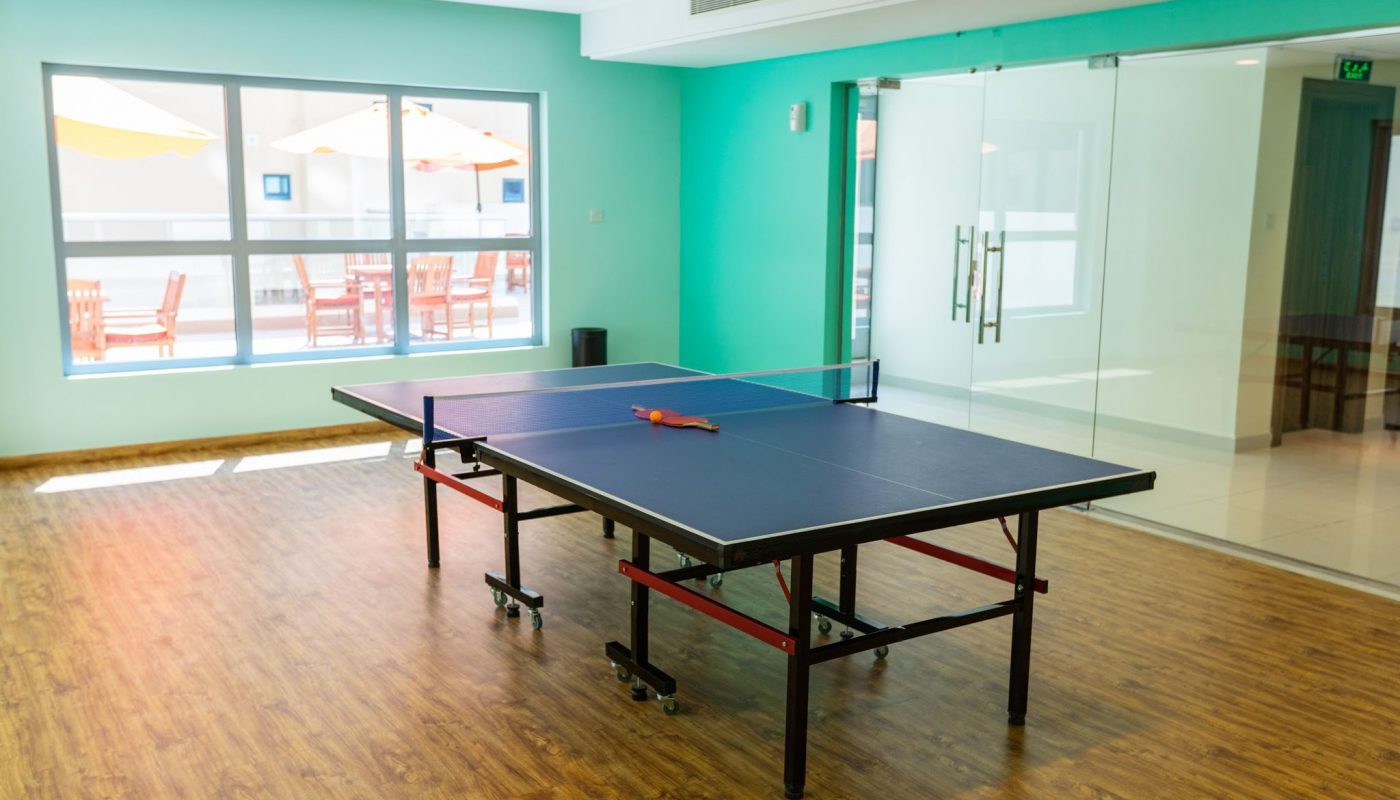 A blue ping pong table with paddles and a ball on top, set in a bright room with wood flooring, large windows, and glass doors.