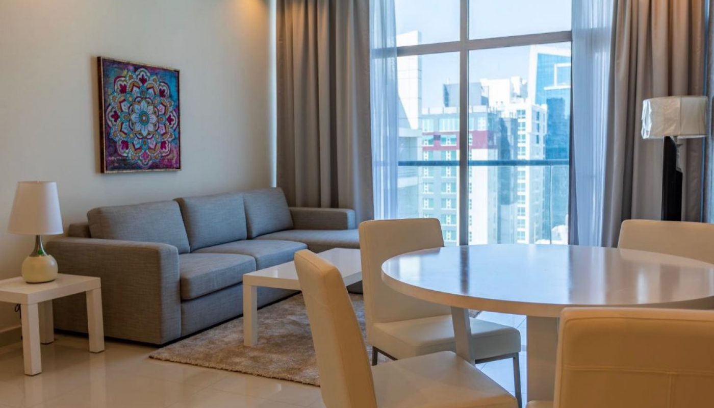Modern living room with a grey sofa, white side tables, a round dining table with four chairs, large window, and city buildings visible outside.