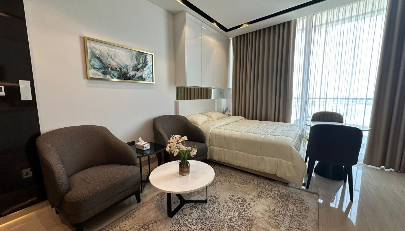 Modern hotel room with a bed, two armchairs, a small round table with flowers, a rug, a desk and chair, large window with curtains, and a wall painting above the bed.