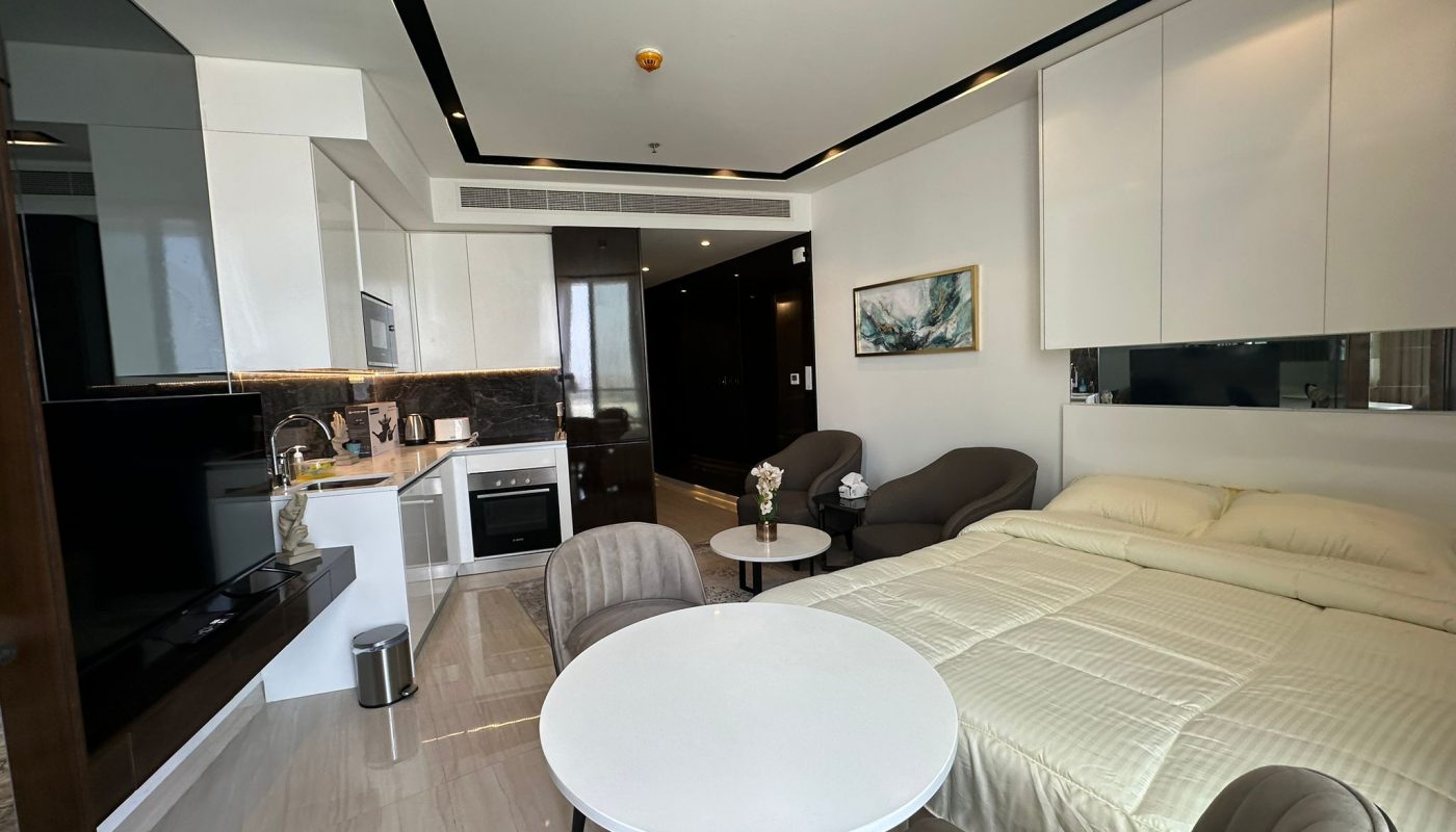 Modern studio apartment with a bed, small dining table, chairs, kitchenette, TV, and neutral decor.