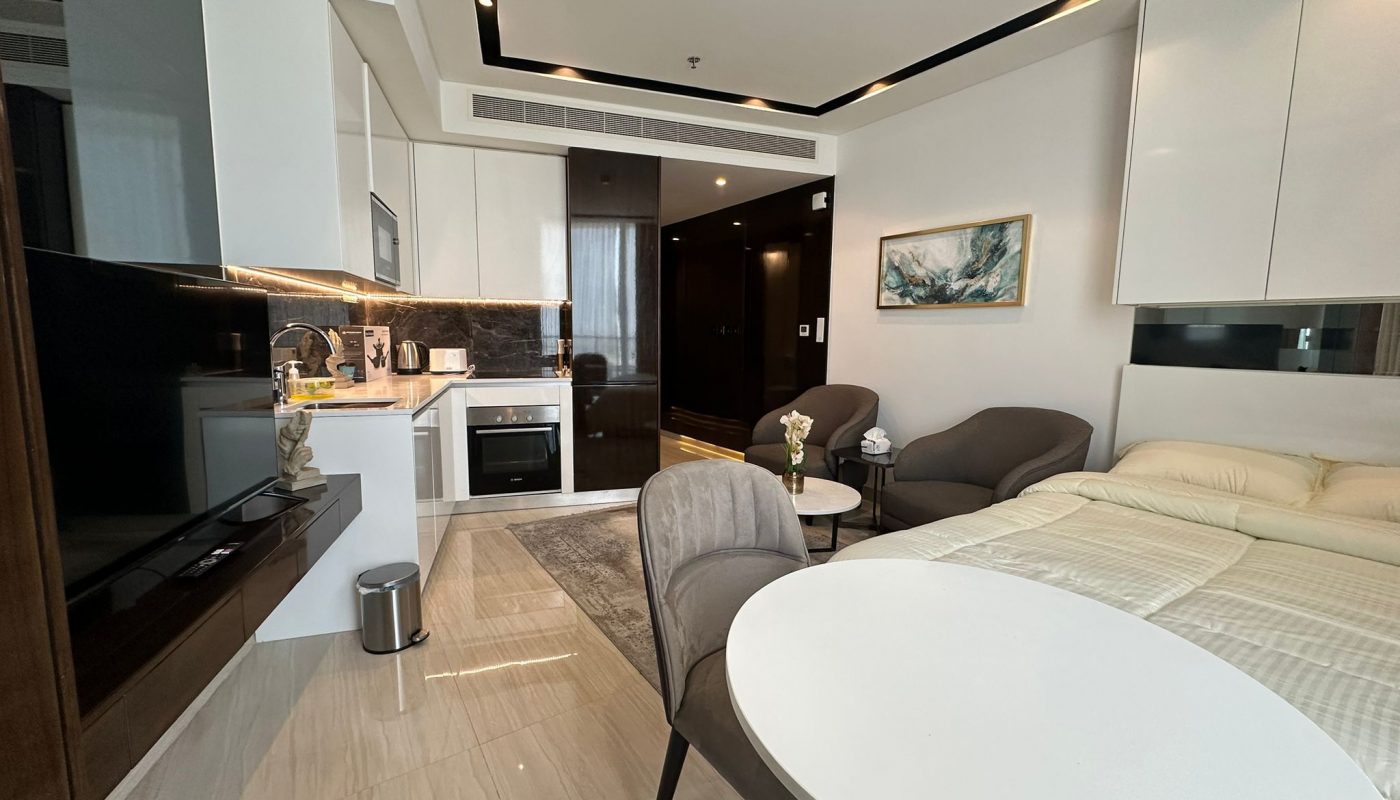 Modern studio apartment with a kitchenette, round dining table, chairs, bed, and seating area; neutral color palette and contemporary decor.