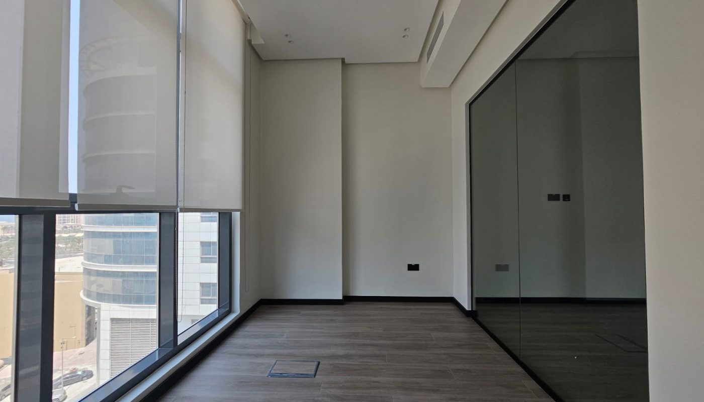 Empty office room with large windows, roller blinds, wood flooring, white walls, and a glass partition reflecting part of the space.