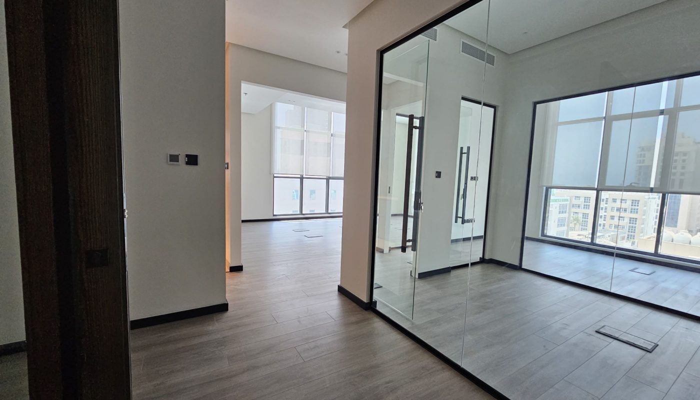 A modern, unfurnished office space with large windows, glass partitions, and wood-look flooring, featuring views of city buildings outside.