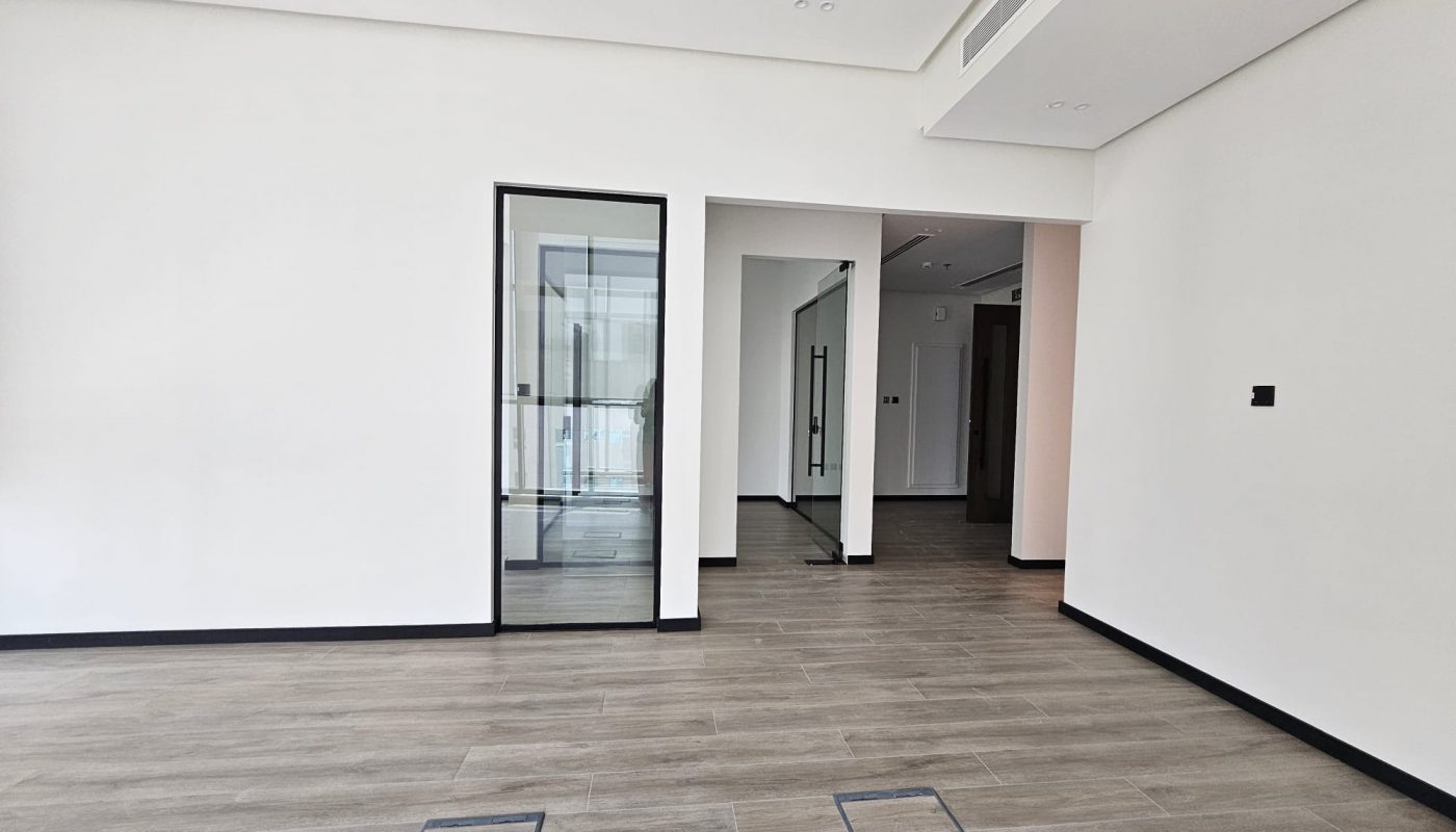 A modern, empty room with light wood flooring, white walls, glass door, and recessed ceiling lights.