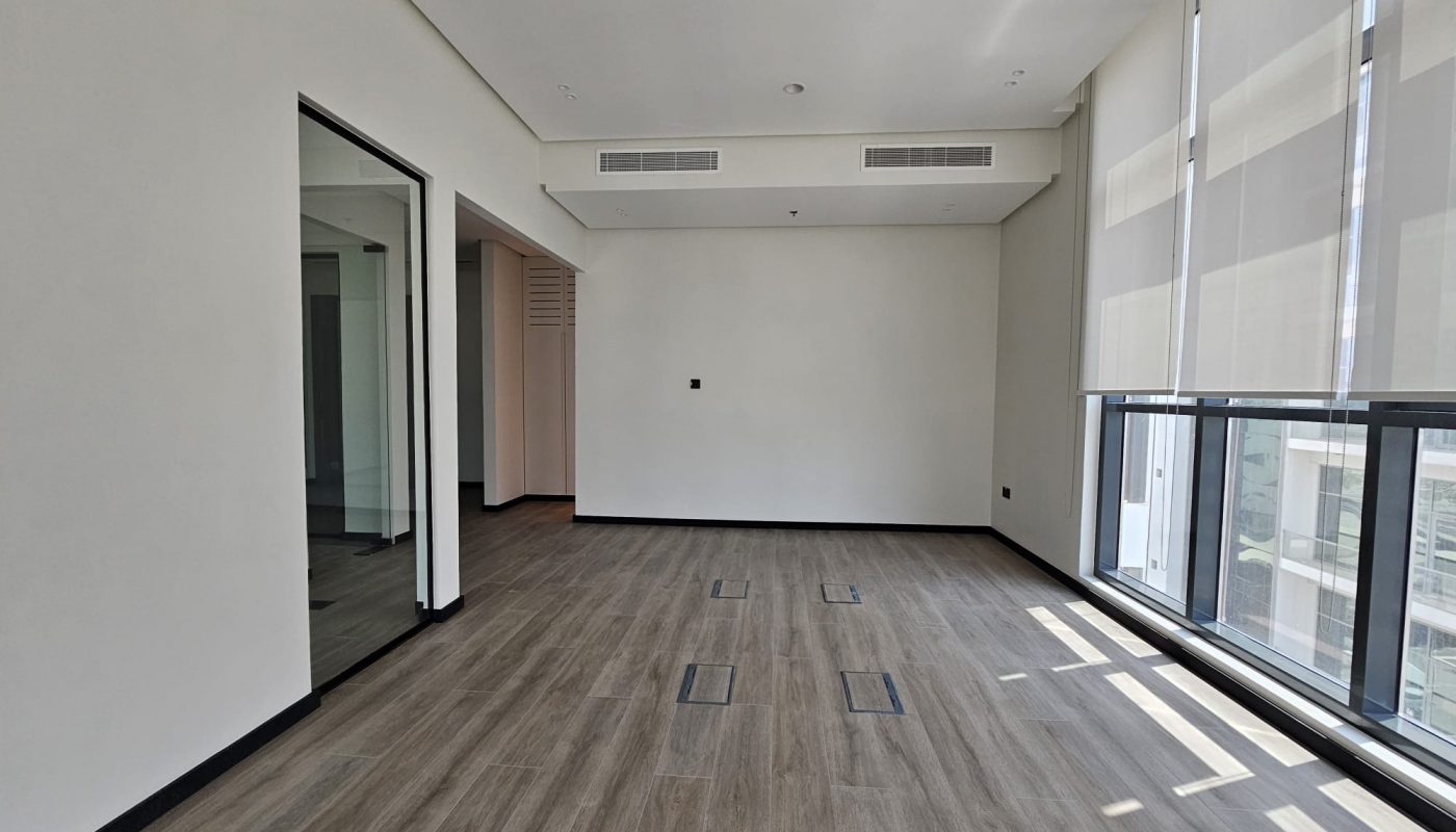 Unfurnished office space with wood flooring, large windows with blinds, white walls, and ceiling air vents. Natural light brightens the room.