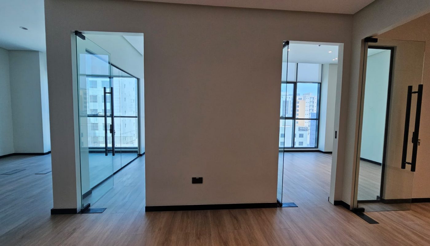 Modern office space with glass-walled rooms, wooden flooring, and large windows showing city buildings in the background.