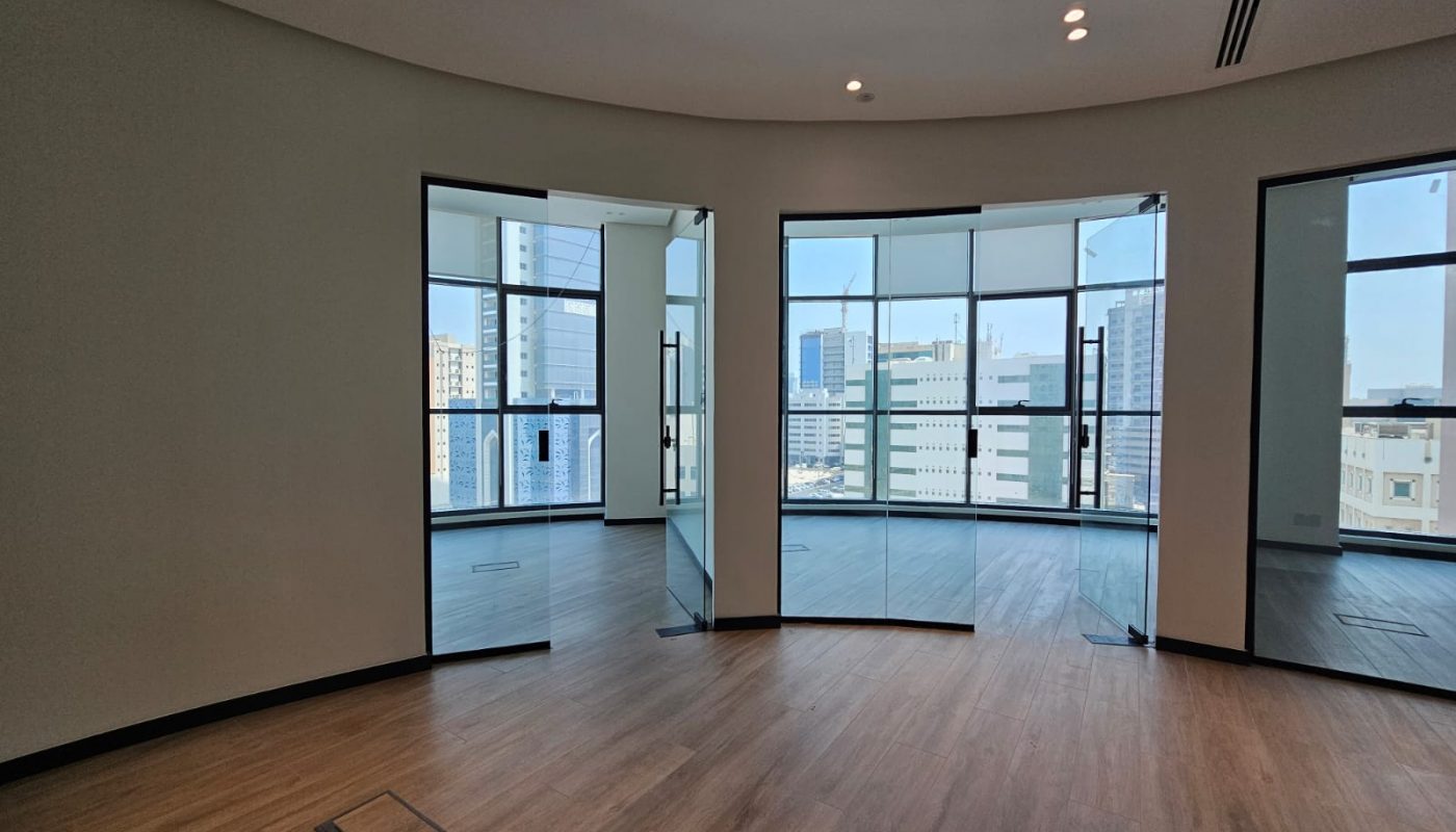 Empty modern office space with wood flooring, large curved windows, and glass doors, offering a view of multiple city buildings outside.