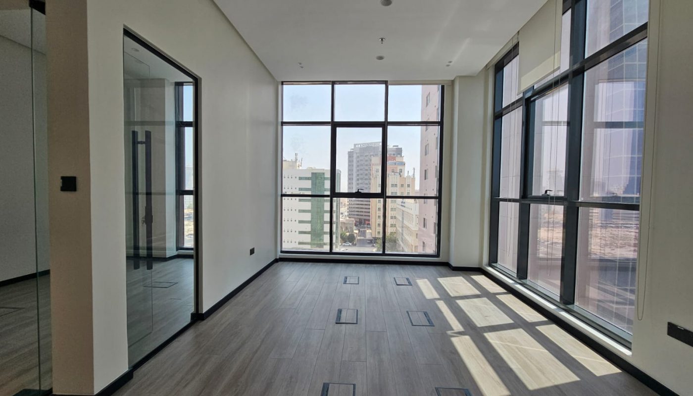 Modern empty office space with large floor-to-ceiling windows, wood-style flooring, and city buildings visible outside. Natural light fills the room.