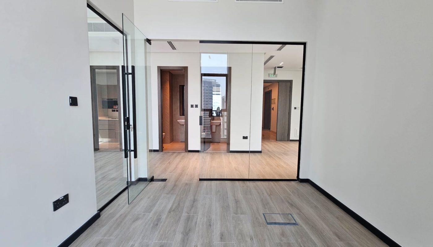 A modern, unfurnished office space with glass doors, mirrored wall panels, wood flooring, and neutral-colored walls.