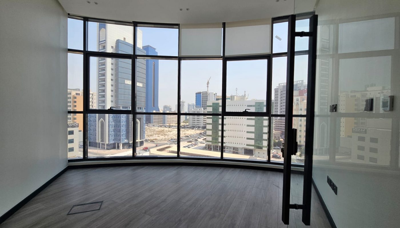 Modern, empty office room with large floor-to-ceiling windows overlooking a cityscape with tall buildings and clear skies.