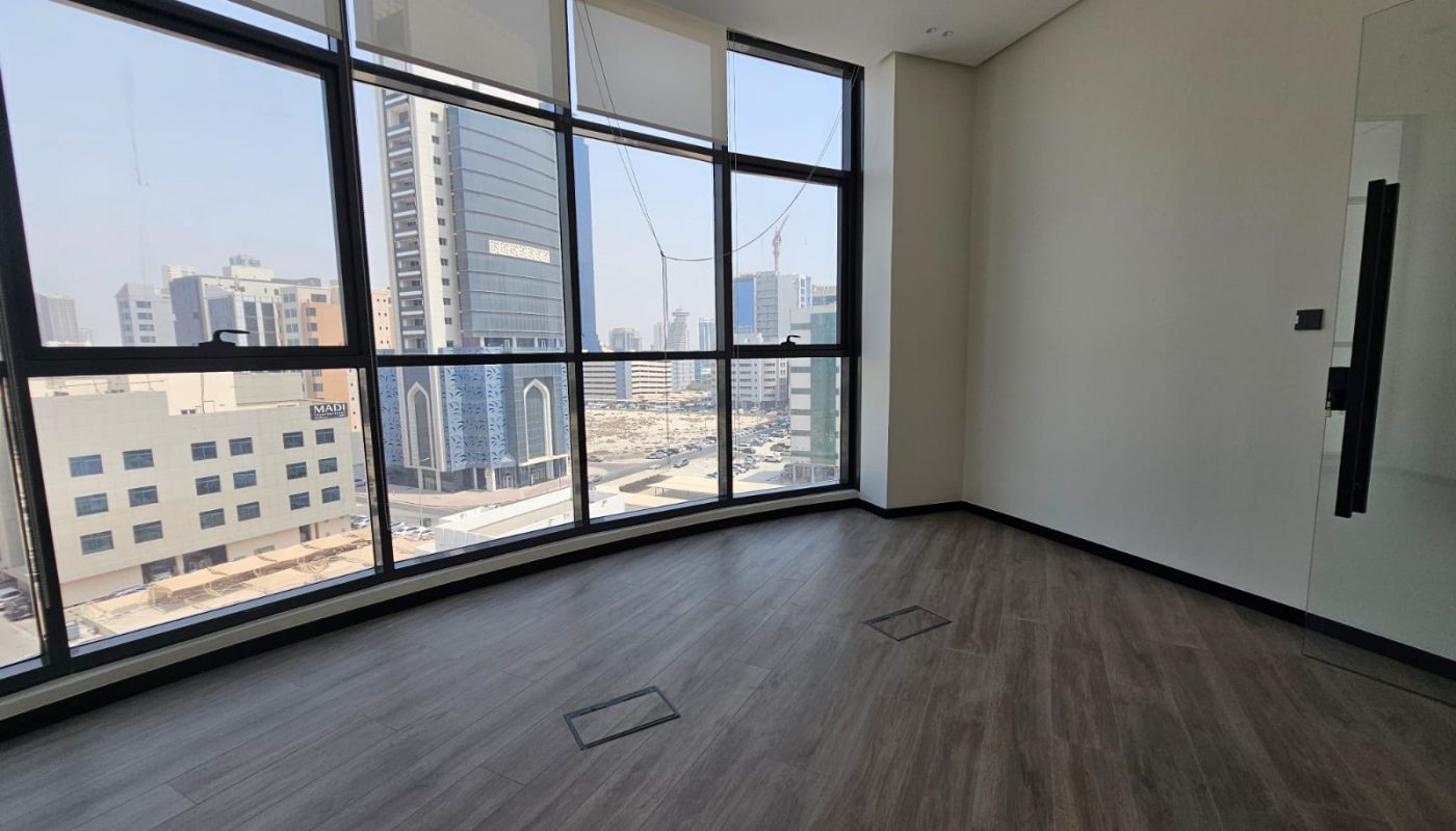 Empty office room with large floor-to-ceiling windows, wood-look flooring, and a view of city buildings outside during daytime.