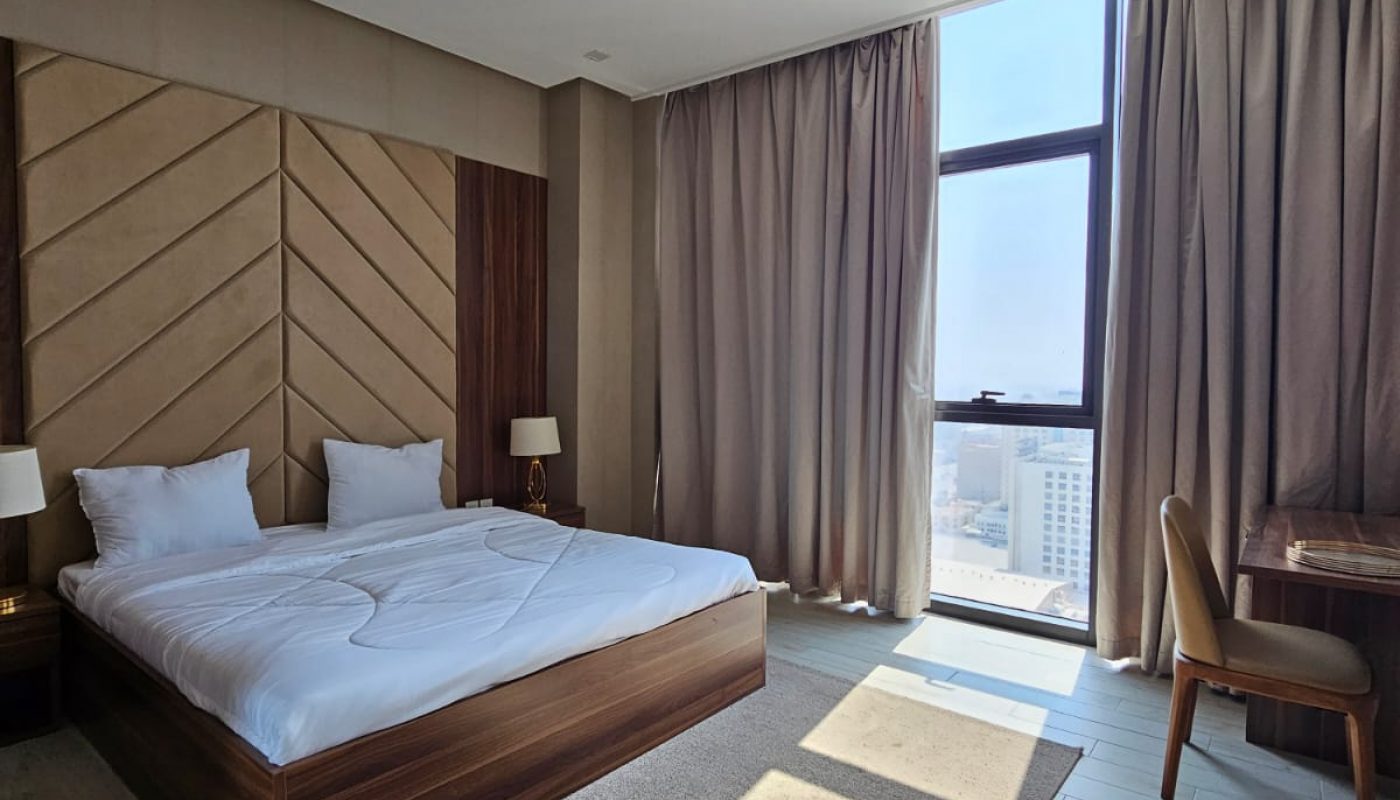 Modern hotel room with a large bed, two bedside tables, a desk and chair, floor-length curtains, and a window showing a city view.