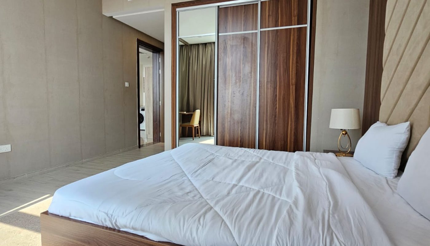 A modern bedroom with a neatly made bed, wooden sliding closet doors, a lamp on a side table, and a chair visible through the open door in the background.