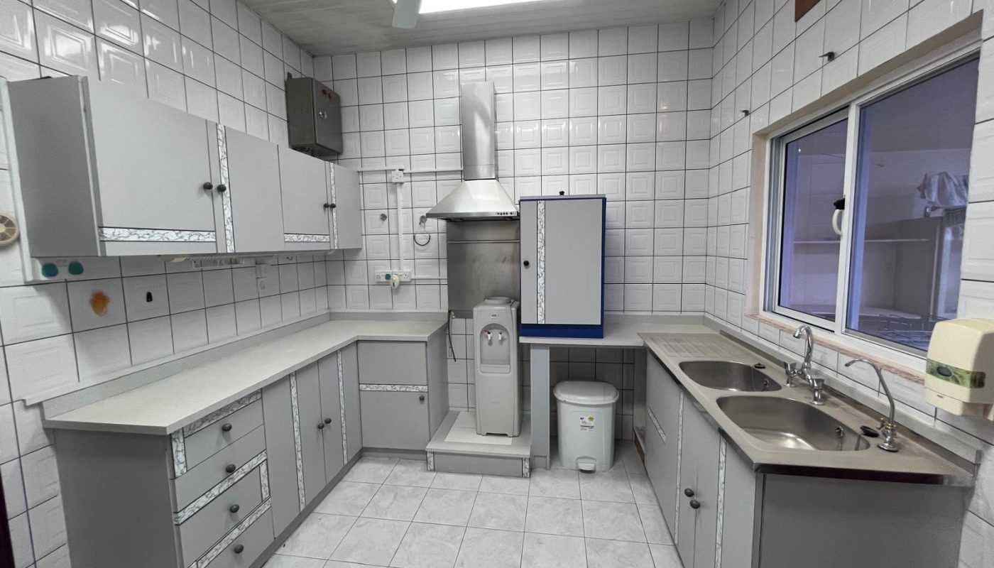 A clean, tiled kitchen with grey cabinets, dual sinks, wall-mounted cabinets, a stove with hood, and a large window above the counter.