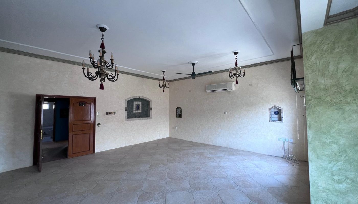 Empty room with tiled floor, cream-colored walls, two chandeliers, ceiling fans, air conditioning unit, and an open wooden door. Decorative wall accents and light fixtures are visible.