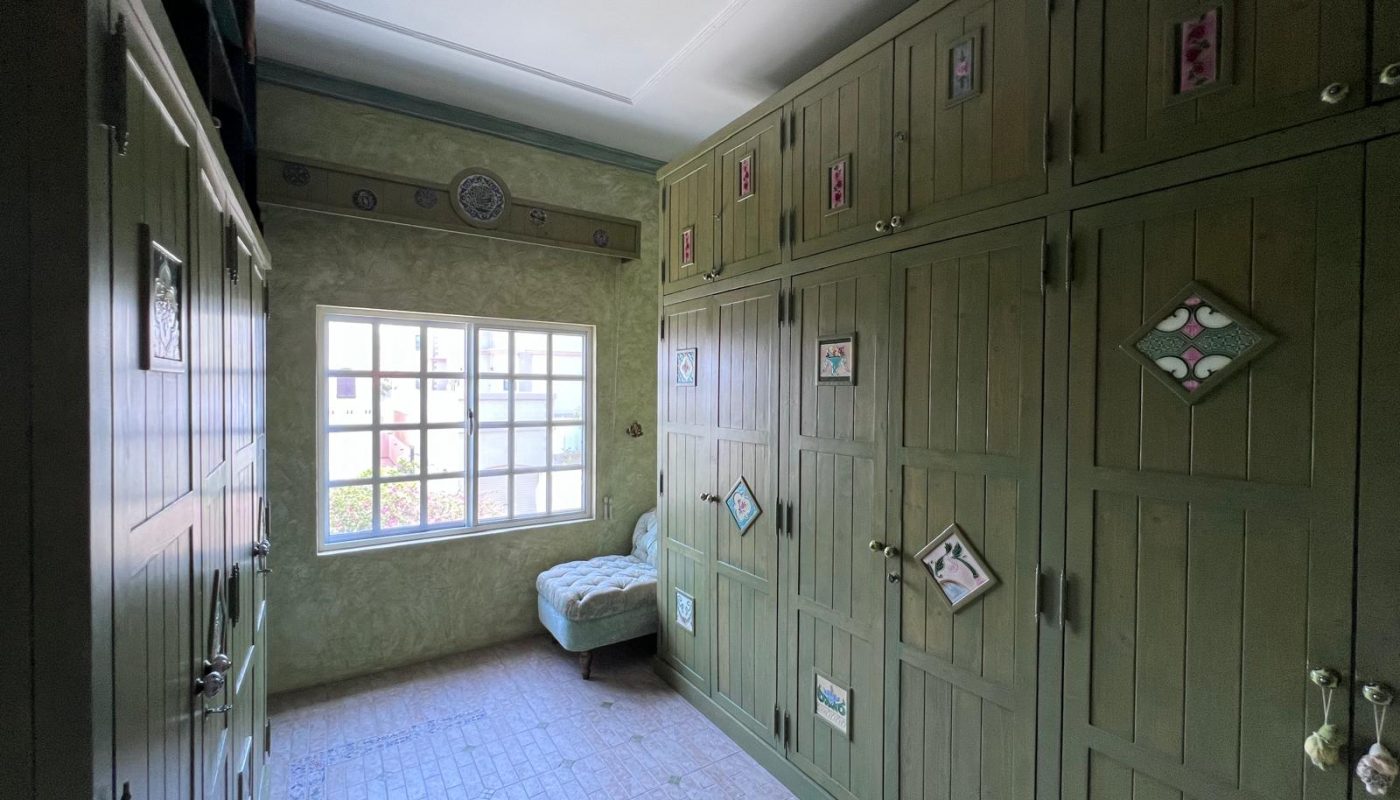 A room with green built-in wardrobes, a tiled floor, a window letting in natural light, and a light green upholstered bench.