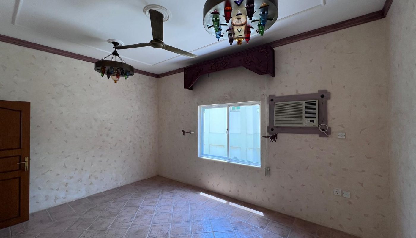 Empty room with tiled floor, beige patterned walls, ceiling fan, two decorative light fixtures, a closed wooden door, a window, and a wall-mounted air conditioning unit.