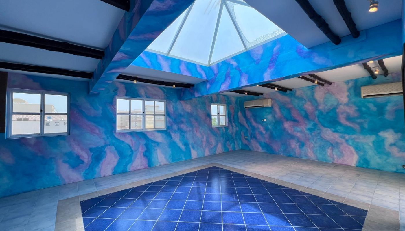 Room with blue and purple cloud-patterned walls, a glass ceiling, wooden beams, two windows, and a central blue tiled floor area.