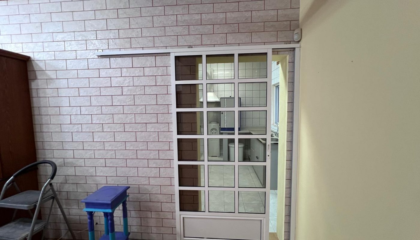 Sliding glass door with a white grid frame separates two rooms; a purple step stool and a folding ladder are placed nearby against a tiled wall.
