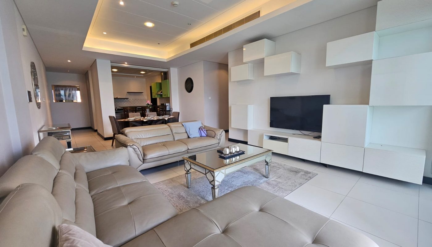 Modern living room with beige sectional sofas, glass coffee table, wall-mounted shelves, flat-screen TV, and open view into the kitchen and dining area.