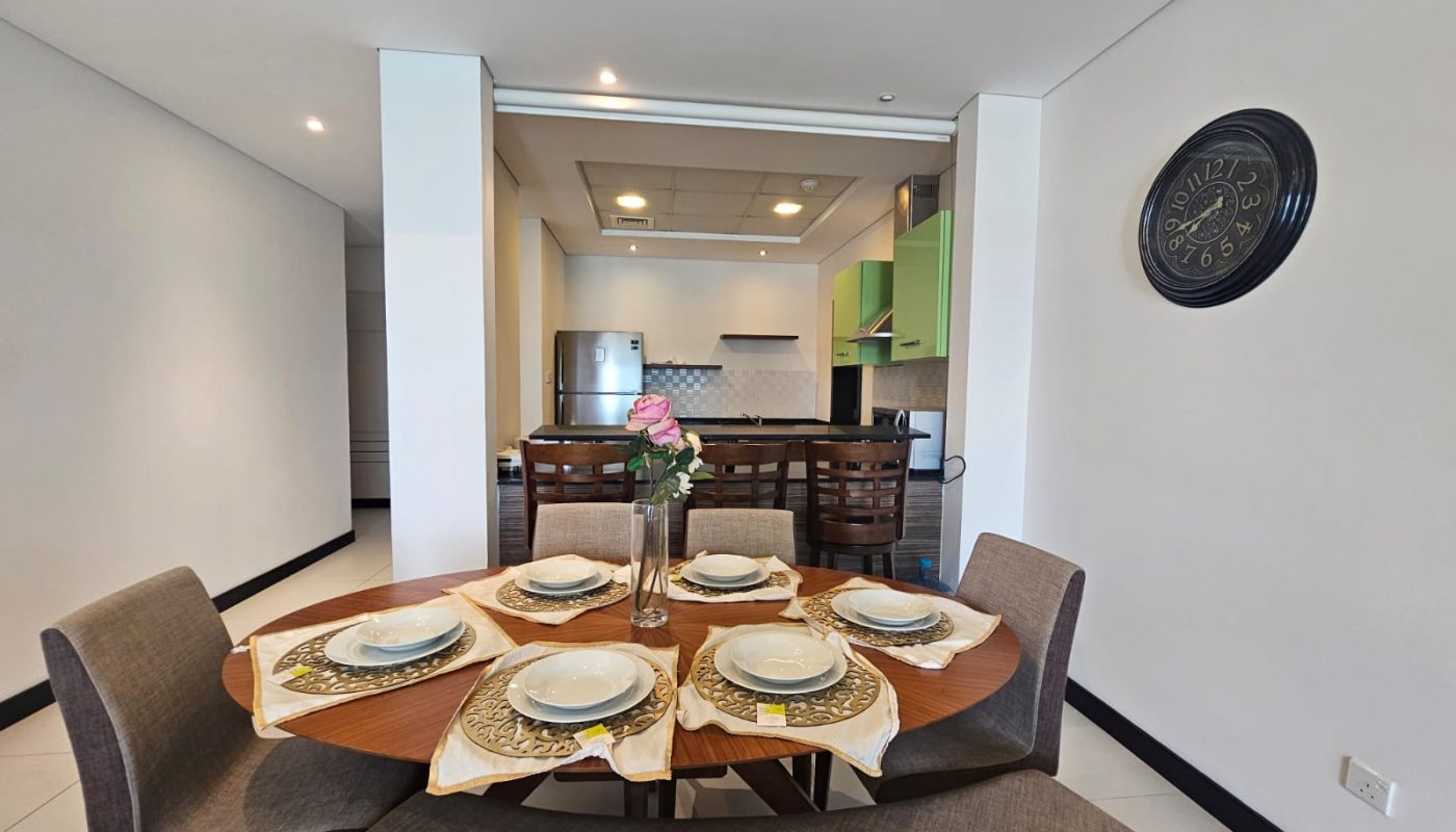 A modern dining area with a round wooden table set for four, adjacent to an open kitchen with a refrigerator, stools, and a wall clock.