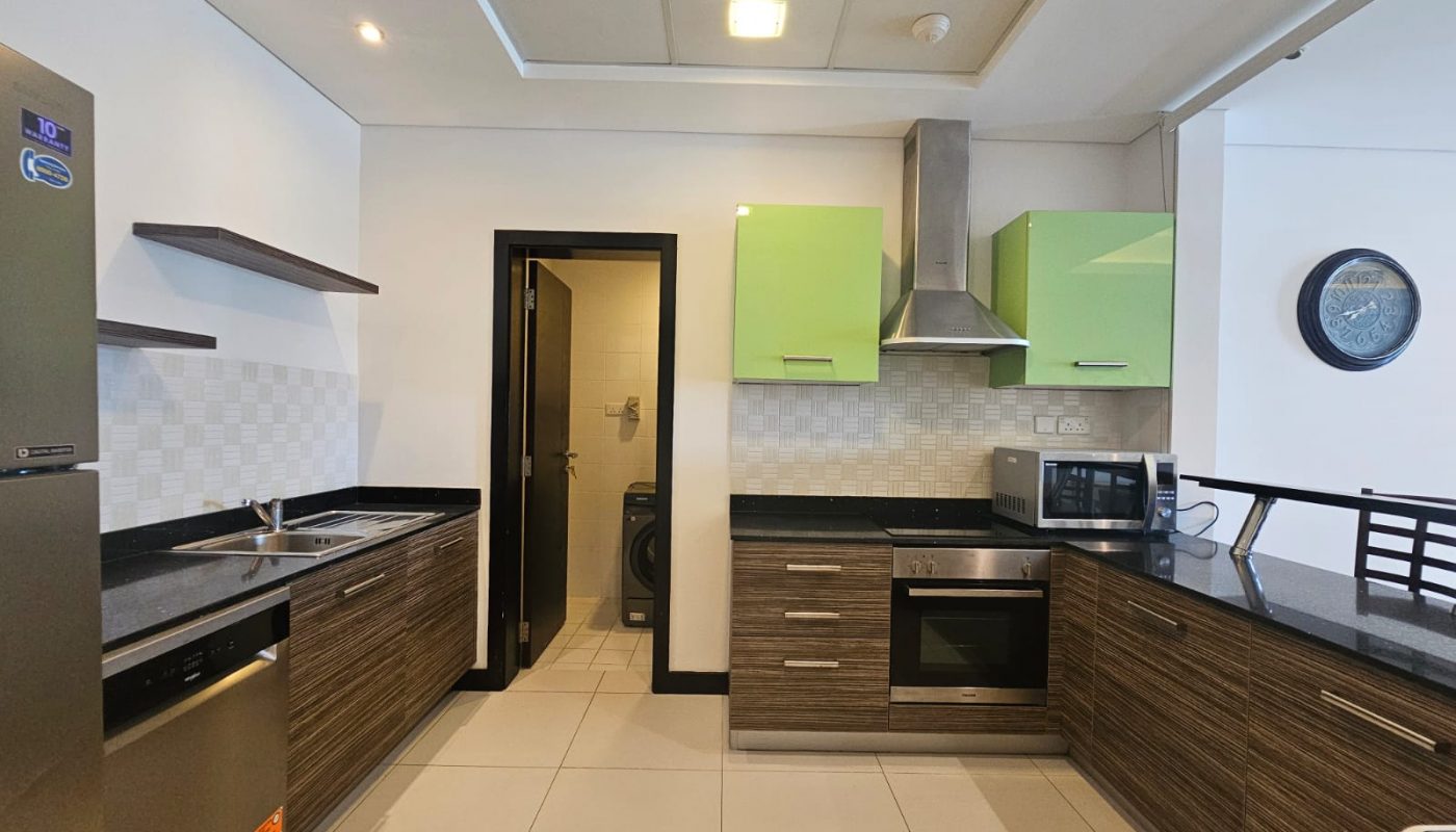 Modern kitchen with green upper cabinets, wood-textured lower cabinets, built-in oven, microwave, refrigerator, dishwasher, and a visible laundry area through an open door.